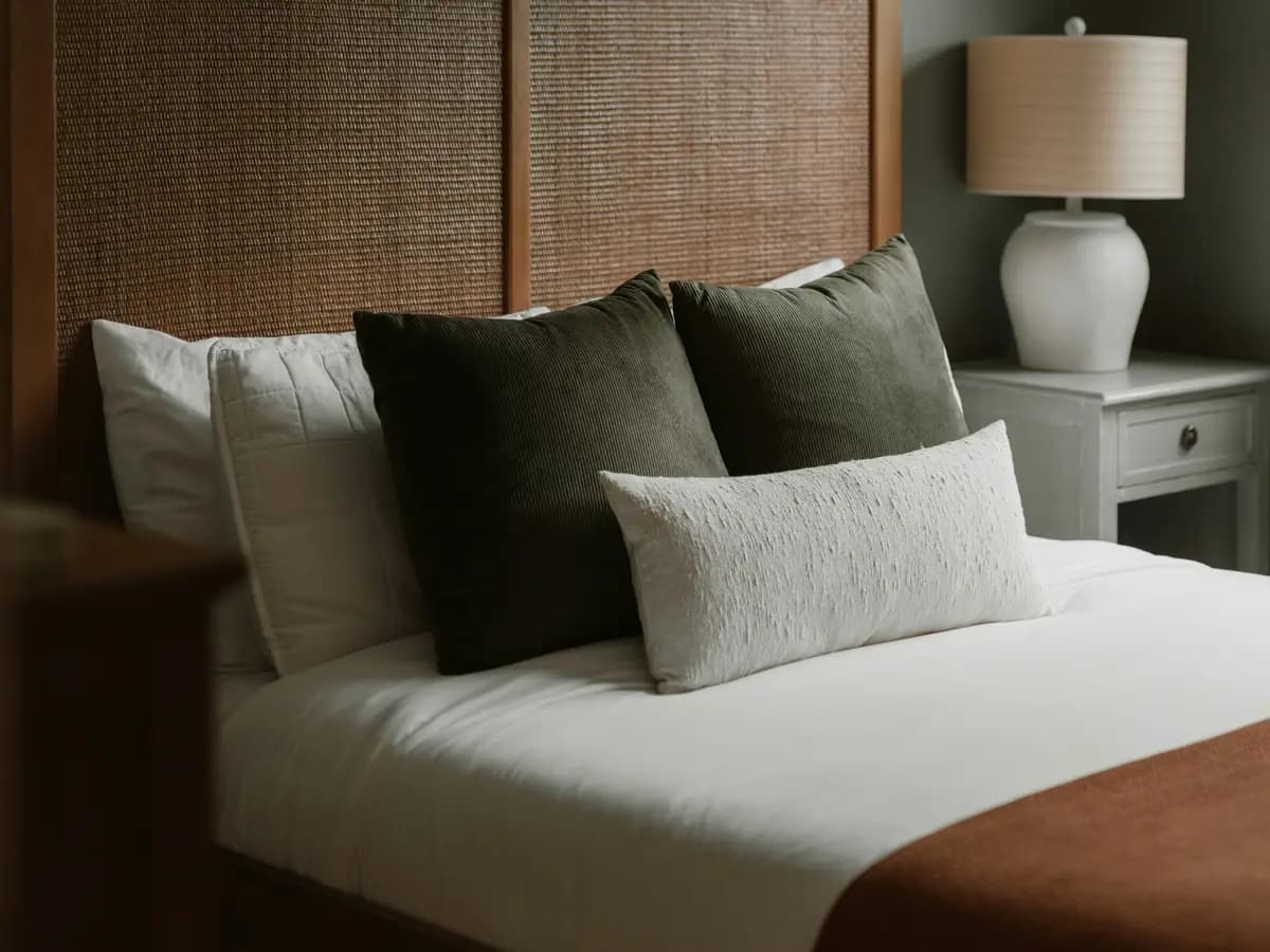 Neatly made bed with layered pillows, a woven headboard, and a bedside lamp.